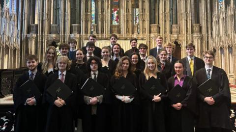 Group of students standing in a church in black gowns holding choir books with their hands folded.