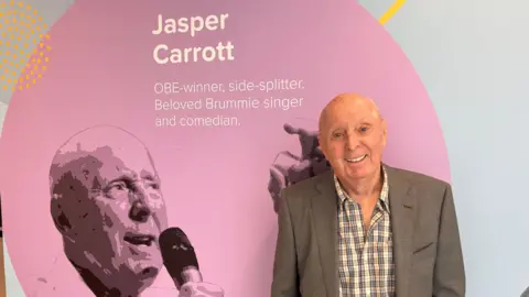 Jasper Carrott stood against a purple and light blue wall with his face printed on it alongside words that read "OBE-winner, side spiller. Beloved Brummie singer and comedian. He is stood next to it smiling in a grey blazer jacket and checked shirt