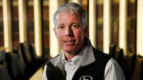 Getty Images Chris Wright, who has short grey hair and is wearing a black quarter-zip vest over a white shirt, poses for the camera against a boardroom backdrop with neon lights on the wall behind him