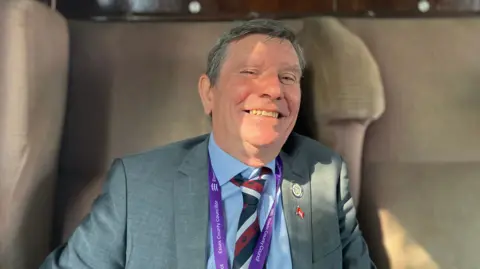 Henry Godfrey-Evans/BBC Jeff Bray has greying dark hair and is wearing a grey suit, blue shirt and striped tie. He is smiling widely at the camera and is sitting in a beige train seat. 