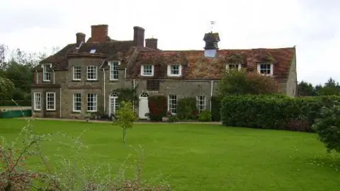 Phil Catterall/Geograph A large, traditional stone country house with a red-tiled, weathered roof sits behind a wide, neatly mown lawn. The building has multiple chimneys, white-framed windows and a central doorway, with sections of ivy or climbing plants visible on parts of the walls. In the foreground, a shrub with thin branches and small red berries spreads across the grass. Hedges and bushes border the lawn, and trees line the background under an overcast sky.