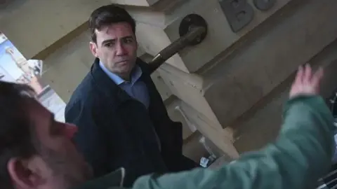 GMCA A young looking Andy Burnham looks on as a blurred man out of shot gesticulates with his hands