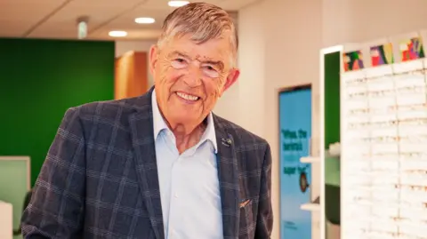 Man in a jacket which is blue and white and a light blue shirt. He has grey and white hair and is in a Specsavers store