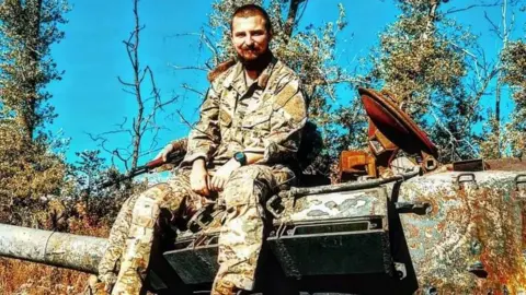 Family handout A soldier sitting on a tank. He has shaved brown hair and is smiling. He's wearing khakis and a watch. The sky is bright blue and cloudless. There are trees surrounding the tank.