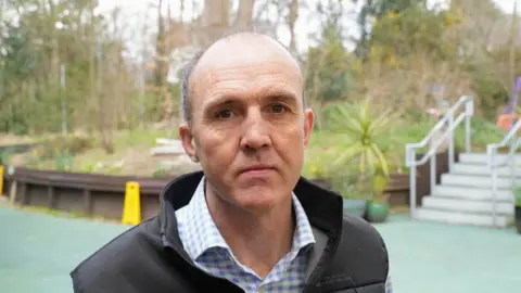 Ian Nicolson wearing a blue checked shirt and a black gilet. He has short grey hair and is cleanly shaven. He is stood in front of a garden