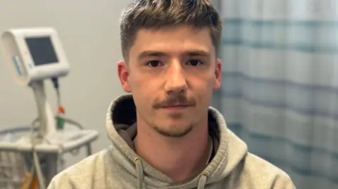 A man with dark hair, short and swept to his right side, looks in the camera. He has a short moustache and chin beard. He wears a grey hoodie top with the hood pulled back. Behind him is a blue checked curtain and on the other side, a monitor on a stand.