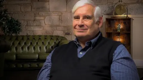 IWC Media Mick North is a white-haired man in a blue sleeveless pullover and checked blue shirt. He is smiling in front of a stone wall, green leather sofa and lit cabinet full of ornaments.