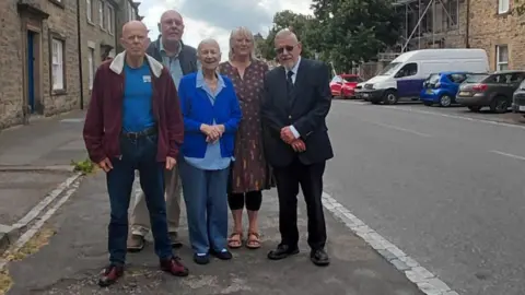 Five people stand in a group next to the road. Chris Foote-Wood is on the right with Andrew Moorhouse and Jo Angell at the front. Pete Whittaker and Ros Evans are at the rear. The road is bordered by white stone terraced houses. There is a line of vehicles in a carpark on the far right.