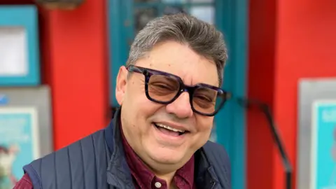 Ugur Vata smiles at the camera outside his restaurant. He has short grey and dark hair and he is wearing tinted glasses with black rims. He is wearing a navy gillet with a red shirt underneath. The restaurant behind him has a bright red exterior with a teal coloured door.