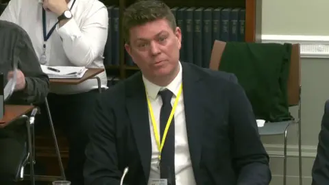 NI Assembly A man wearing a suit and tie and yellow lanyard sitting at a desk in a Stormont committee room. There is a book shelf and some other people behind him.