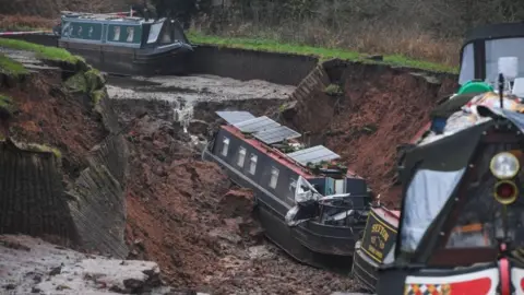 A canal boat in a large muddy hole with other boats seen in a drained trench on either side