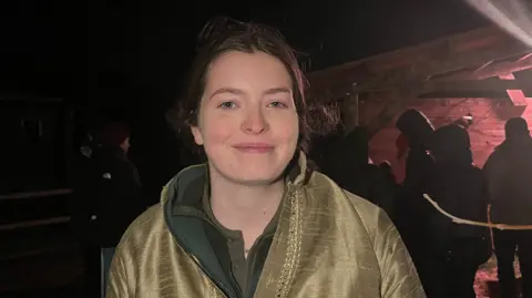 John Devine/BBC Kat Choppin has brown hair tied behind her head and is wearing a golden, embroidered jacket with a dark fleece underneath. Behind her is a stable and a few people near it, in darkness.