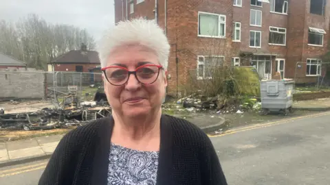Photograph of Francesca Estasy, who lived on Baguley Crescent. The image shows a burnt-out caravan and an abandoned block of flats in the background.