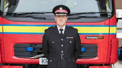 Stuart Hook, dressed in a black fire chief uniform, stands in front of a fire engine. 