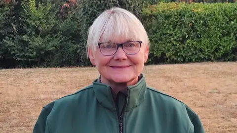 A woman with short blonde hair and glasses looking at the camera. She is wearing a green top with a zip. Light brown grass and a hedge are in the background.