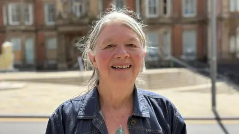 Joan Walley, a woman wearing a denim jacket, is standing outside Hanley Town Hall. She is smiling for the photograph.