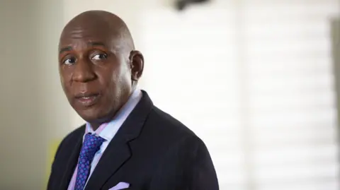 A man stands off centre in a suit and tie. Behind him a blind is blurred. He looks off to the right.