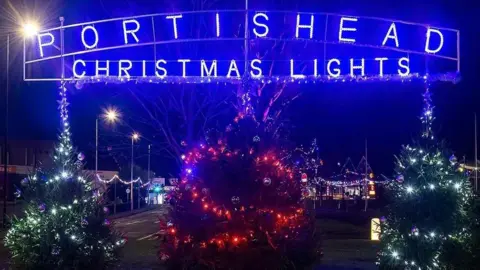 Portishead Christmas Lights - the words are lit up in blue lights above three Christmas trees dressed in fairy lights. Behind the trees the street can be seen with more lights.