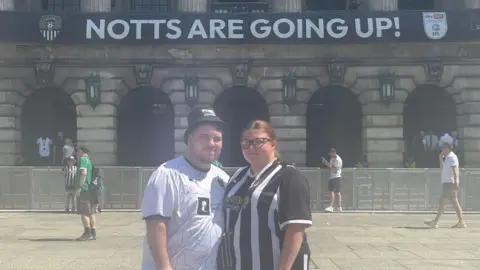 Two people in Notts County shirts in front of the council house with a banner that reads NOTTS ARE GOING UP