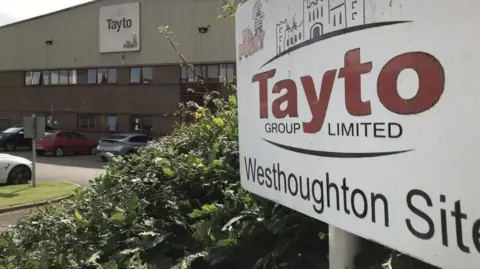 A sign outside a factory says Tayto Group Ltd Westhoughton site, with several cars parked in front of the building.