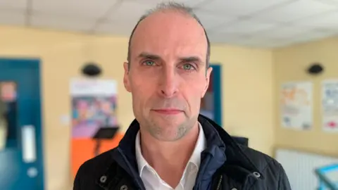 Mark Cairns, he is looking at the camera, he has dark hair and is wearing a white shirt with a navy coat.