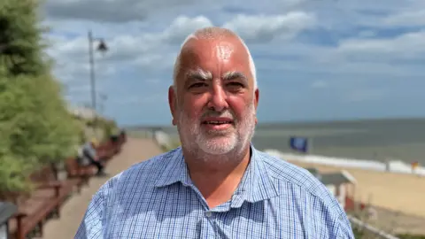 George King/BBC A head and shoulders image of Steven Janes. He is wearing a shirt and smiling into the camera. The Southwold promenade and beach can be seen in the background.
