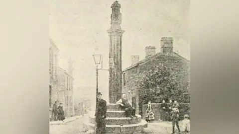 Kirklees Council A black and white photograph of two men standing by a stone cross in Victorian fashion. There are houses to the left and right of the cross