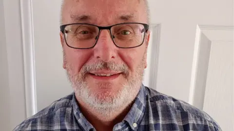 Andy Sturrock is stood against a door and has a short grey beard and dark framed glasses.