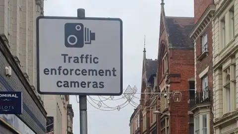 A Traffic enforcement camera sign, in a town centre, with stone and brick buildings, on either side of the sign. There are lights across the road. 