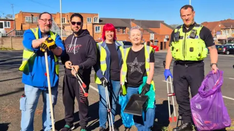Four people smiling holding litter pickers. Three of them are wearing a yellow hi-vis and there is also a police man holding a purple plastic bin bag.