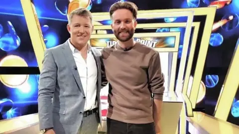 Publicity image of Ben Shephard and Jamie posing together on the Tipping Poin set, which has a blue background and a row of yellow square arches above.