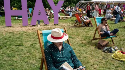 Getty Images A person in a white brimmed hat and shirt is seen sat reading a book, in front of a large purple sign reading "Hay" 