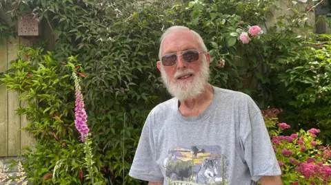 A man with grey hair and glasses with a grey tshirt standing in a garden with pink flowers and a wooden fence