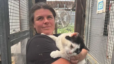 Emma Floyd is standing just off centre wearing a black T-shirt looking at the camera. She is holding a white and black cat in her arms, and is standing in a metal animal cage.