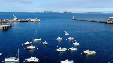 Fourteen white boats float in dark blue water, with two piers and islands in the distance.