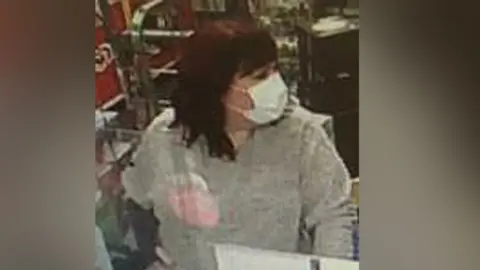 Hertfordshire Constabulary A woman with shoulder-length dark red hair wearing a Covid-style white face mask standing at a till. She is looking to her right and wearing a grey sweater