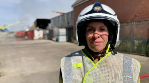 Claire Tait - a woman in a white helmet and a high vis vest - is smiling into the camera. The background is blurred but she is standing in an industrial area.