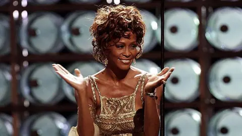 Getty Images Whitney Houston tampil di panggung pada Penghargaan Musik Dunia 2004 di Thomas dan Mack Center, Las Vegas pada tahun 2004. Dia mengenakan gaun bertali emas.