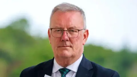 Mike Nesbitt has a neutral expression on his face as he stares at the camera. He has short grey hair, clean shaven and is wearing wired square-framed glasses, a navy suit jacket, a white collared shirt and a navy and green striped tie. He is standing in front of some trees but they are blurred in the background.