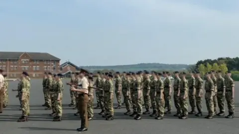 Family Handout A screenshot from a video showing a regiment practicing their uniform marching and saluting on camp at Larkhill. There is a large group of about 30 people dressed in green and brown camouflage uniforms and black caps. They are standing on a large tarmac area surrounded by fields and trees. In the background there is a brown brick building with lots of windows.