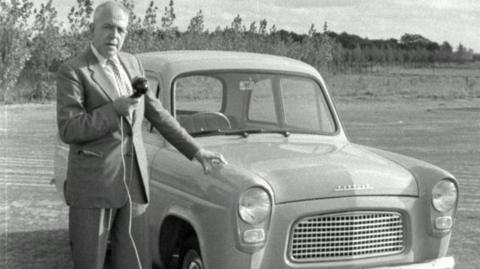 Man stands beside Ford Popular car with handheld microphone.