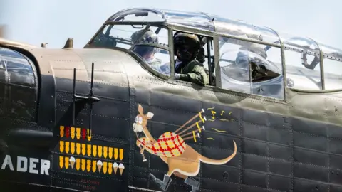 Claire Hartley Three crew-members look out from the glass cockpit of a Lancaster bomber. The plane is painted partly in brown and green camouflage, and partly in black. A logo shows a cartoon kangaroo playing the bagpipes.