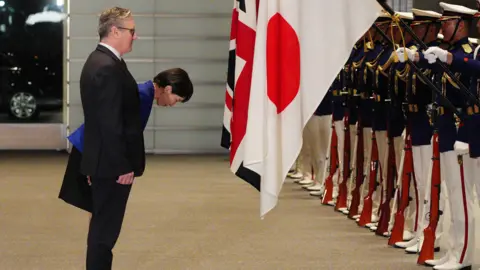Reuters The British PM stands, and Japan's PM bows, in front of guards wearing white and navy unifroms and caps and holding red rifles, and UK and Japanese flags 
