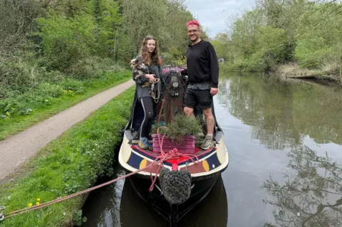 A young man with pink hair and glasses stands on a narrowboat moored on a river next to a young woman with long wavy hair who is holding a small dog. There is a towpath to the left of them. 