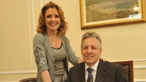PA Media Iris and Peter Robinson smiling in an office. Iris has long red hair and is wearing a black and white top with a cardigan. Peter has short grey hair and is wearing a grey suit with a white shirt and a blue, yellow, dark blue and red striped tie.