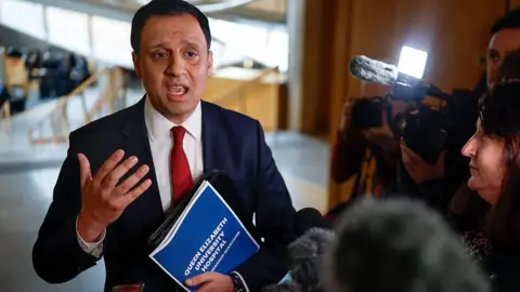 Getty Images Anas Sarwar in blue suit, white shirt and red tie is holding a document bearing the words "Queen Elizabeth University Hospital" and gesticulating while talking to a crowd of journalists. They and their cameras are huddled around him. They are in the lobby of the Scottish Parliament building at Holyrood, which features light wood and grey concrete floors.
