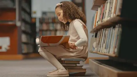 A young girl sits on books in a library and reads. She is wearing a pinafore and tights and a headband and has curly hair.