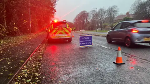 A police car with flashing lights behind a sign for a drink and drug check site. A car approaches and some police officers in high visibility jackets are visible in the background.