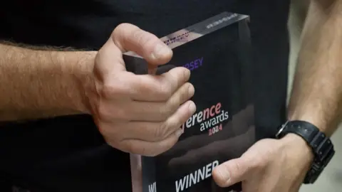 A close up of Jonny's hands as he holds the awards. It's a rectangular glass award with the winner logo. Jonny has his left hand on the top corner of the award with his right hand on the bottom. He has a black watch on his right wrist as well.