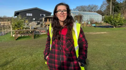 Andrea with her hands in her pockets. She is smiling and wearing glasses. She has got a hi-vis jacket on and a red jacket. The farm can be seen behind her.